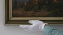 Eine Hand im weißen Handschuh greift nach einem Gemälde in goldenem Rahmen.