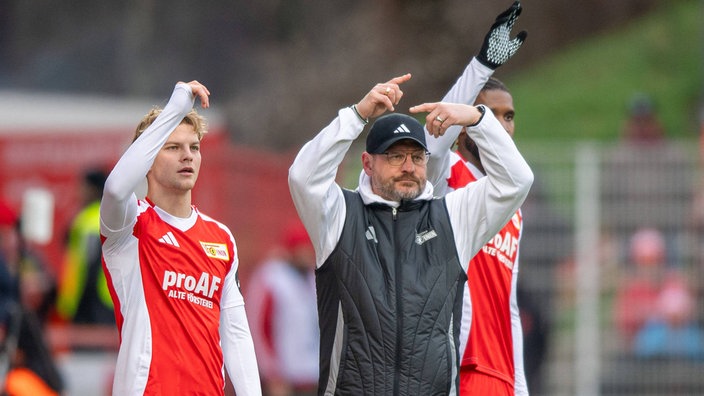 András Schäfer, Trainer Steffen Baumgart und Jordan Siebatcheu von Union Berlin heben nebeneinander am Spielfeldrand die Arme (v.l.n.r.)