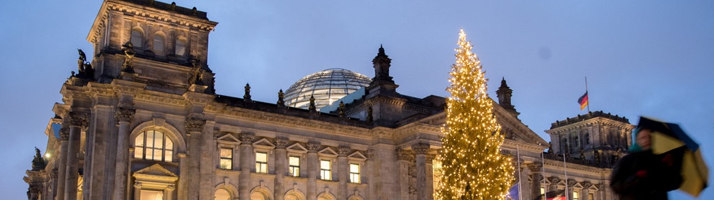 Front des Reichstagsgebäudes in der Dämmerung bei Nässe mit Deutschlandflaggen und mit beleuchtetem Weihnachtsbaum im Vordergrund