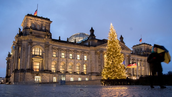 Front des Reichstagsgebäudes in der Dämmerung bei Nässe mit Deutschlandflaggen und mit beleuchtetem Weihnachtsbaum im Vordergrund