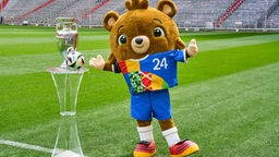 Lebensgroßer Teddybär auf Stadionrasen in buntem Trikot und mit schwarz-rot-goldenen Schuhen neben dem Pokal und dem Spielball der UEFA EURO 2024