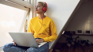 Junge Frau mit Ohrhörern sitzt am Laptop auf der Fensterbank