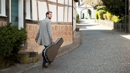 Der Künstler Chris Weule geht mit einem Gitarrenkoffer einen Weg vor einem Fachwerkhaus entlang.