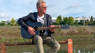 Der Künstler Frank Heidemann sitzt mit seiner Gitarre auf einem Zaun