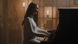 In einem spärlich beleuchteten Raum sitzt Hannah Stienen in einem weißen Kleid und spielt am Klavier.