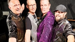 Die vier Bandmitglieder tragen glitzernde Paillettenhemden und stehen vor einem dunklen Hintergrund.