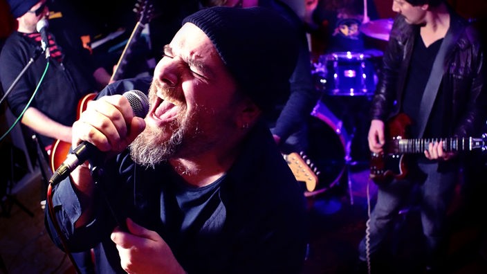 Der Frontmann singt energisch in ein Mikrofon, während im Hintergrund des in Rot- und Blautönen ausgeleuteten Raumes der Rest der Band zu sehen ist.