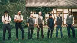 Die 7 Bandmitglieder stehen auf einer Wiese vor einem Haus.