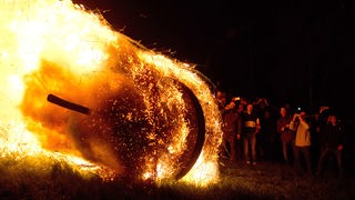 Ein brennendes Eichenrad rollt im Dunkeln an einer Menschenmenge vorbei einen Berg hinunter