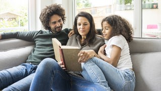 Vater, Mutter und Tochter sitzen mit einem Buch gemeinsam auf der Couch
