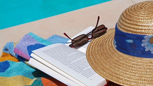 Ein aufgeschlagenes Buch, Sonnenbrille und Strohhut liegen an einem Pool