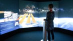 virtuelles Arbeiten in der Zukunft: eine Frau steht vor einer großen Leinwand und hält einen Joystick in der Hand