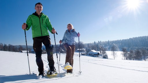 Ein Mann und eine Frau mit Schneeschuhen und Skistöcken laufen bei Sonnenschein durch den Schnee. 