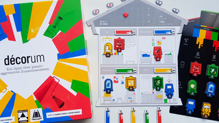 Die Spieleschachtel von "Décorum", daneben ein weißer Hausspielplan mit bunten Einrichtungsgegenständen und Wandfarben und ein schwarzer Spielplan für die verschiedenen Möbvelstücke