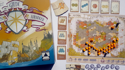 Die Spielschachtel von "Die Gilde der fahrenden Händler" zeigt ein Schiff auf See. Daneben liegt eine Landkarte, auf der sechseckige Felder mit bunten Klötzchen markiert sind.