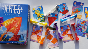 Die Schachtel von "Kites" neben bunten Spielkarten, die verschiedene Lenkdrachen zeigen, und farbigen Sanduhren auf einem Tisch.