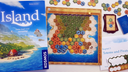 Die Spielschachte von "My Island" auf einem Tisch, daneben ein Spielbrett, dass eine Insel aus sechseckigen Feldern zeigt, auf die Landschaftsplättchen gelegt werden, sowie Kartenstapel und ein Umschlag mit der Aufschrift "Kapitel 3 - Totems und Piraten" 