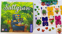 Die Spieleschachte von "Sattgrün" zeigt einen gemütlichen Sessel inmitten wuchernder grüner Pflanzen, daneben das Spielmaterial aus farbenfrohen Spielkarten, Einrichtungsgegenständen und kleinen Blättern aus Holz