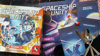 Die Spieleschachtel von Spaceship Unity vor Weltraumhintergrund. Auf diesem sind mehrere Storybücher und Systemkarten angeordnet, dazu Sanduhren, ein paar Socken, Besteck und ein Geschirrtuch