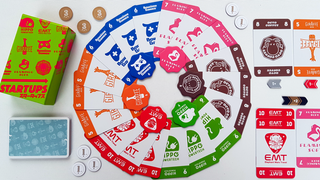Spielschachtel und bunte Spielkarten von "Startups" liegen ringförmig auf einem Spieltisch, daneben Spielchips und weitere Karten