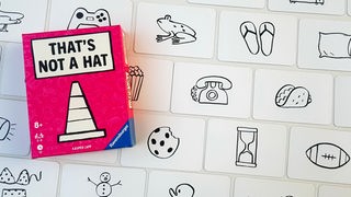 Die Spielschachtel von "That's Not A Hat" zeigt einen Verkehrspylon auf pinkem Hintergrund, daneben Spielkarten mit vielen einfach gezeichneten Motiven wie einem Frosch, einem Keks und einem Telefon