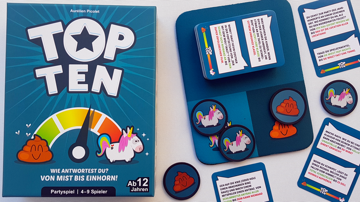 Die Spieleschachtel von "Top Ten" liegt neben einer Spielmatte aus Neopren, auf der ein Kartenstapel und Pokerchips mit Einhorn- und Kothaufen-Motiven liegen