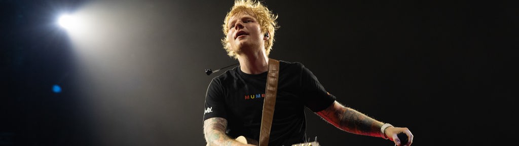 Ed Sheeran steht mit einer Gitarre um die Schulter geschnallt auf der Bühne vor einem dunklen Hintergrund.