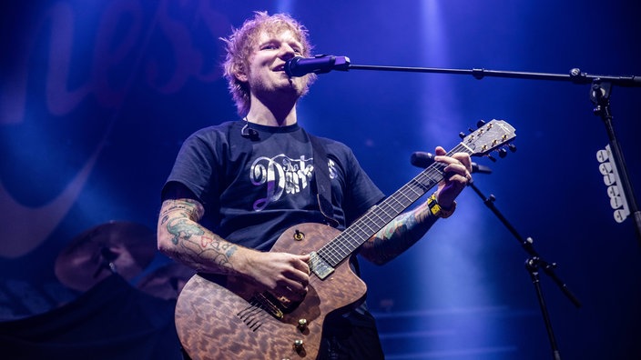 Sänger steht mit Gitarre und Mikro auf der Bühne in blauem Licht.