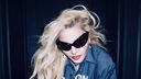 Madonna steht leicht nach vorne gebeugt vor einem dunkelblauen Hintergrund und trägt eine große, dunkle Sonnenbrille.