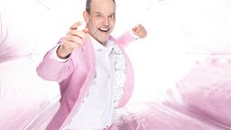 Comedian steht lächelnd vor einem pink-weissem Hintergrund mit pinkem Blazer.