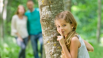 Mädchen versteckt sich hinter einem Baum, Eltern sieht man verschwommen im Hintergrund