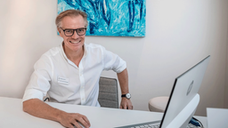 Enneper sitzt in weißer Kleidung an einem Laptop. An der Wand im Hintergrund hängt ein Kunstwerk in Blautönen.