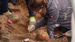 Eine Archäologin legt mit einem Pinsel menschliche Skelette frei. 