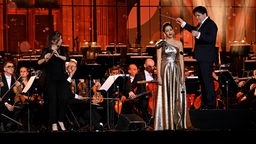 Die Sängerin steht im Goldenen Kleid auf der Bühne. Recht von ihr dirigiert ein Mann, links spielt eine Frau Flöte. Im Hintergrund ist das Orchester.