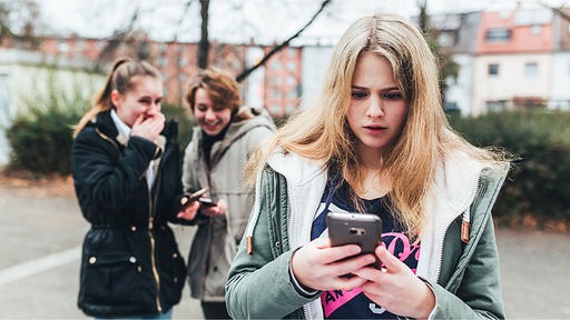 Ein Mädchen schaut schockiert auf ihr Smartphone, während sie im Hintergrund ausgelacht wird. 
