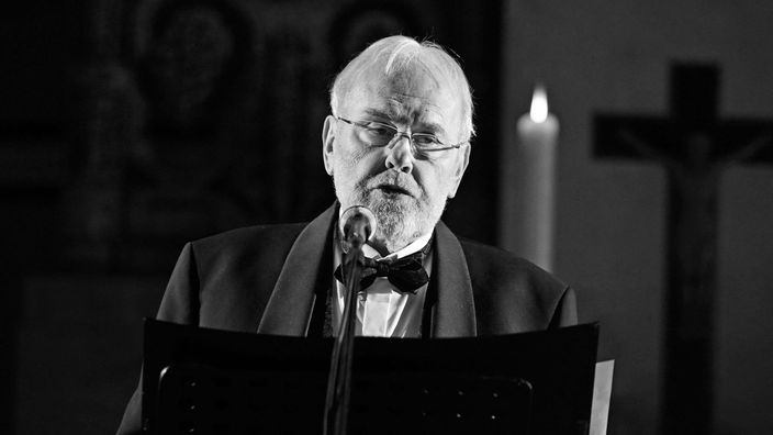 Schwarz-Weiß-Foto von Gunther Emmerlich in festlichen Anzug bei einem Adventskonzert vor einem Mikrofon.