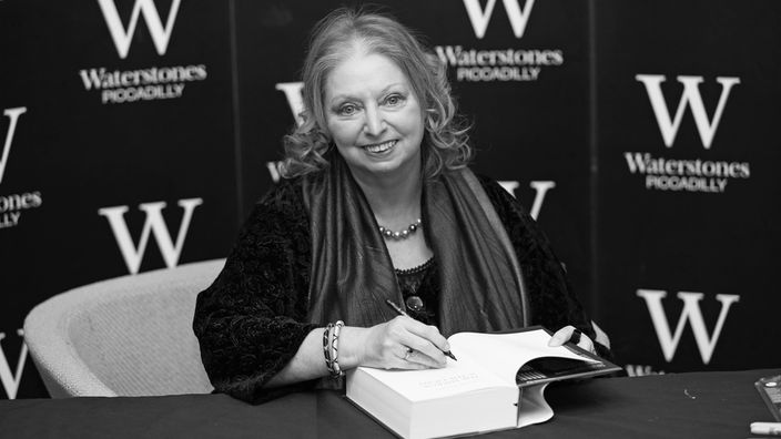 Hilary Mantel sitzt bei einer Buchpräsentation mit einem Stift in der Hand vor einem aufgeschlagenen Buch.
