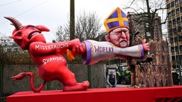 Ein Mottowagen befasst sich mit dem Missbrauchsskandal in der katholischen Kirche und zeigt den Teufel und den Kölner Kardinal Woelki.