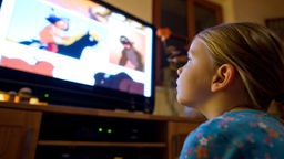 Ein fünfjähriges Mädchen sitzt auf dem Teppich im Wohnzimmer und schaut einen Trickfilm im Fernseher.