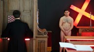 Nackte Männer und ein Mann in einem schwarzen Talar agieren bei einer Kunstaktion. Bei der Inszenierung des Bonner Künstlers Dennis Josef Meseg auf der Discovery Art Fair Köln inszenieren sechs Aktmodelle "lebende Gemälde". 