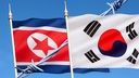 Eine nordkoreanische und eine südkoreanische Flagge nebeneinander gehisst, dazwischen befindet sich ein Stacheldraht.