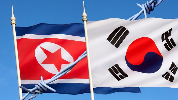 Eine nordkoreanische und eine südkoreanische Flagge nebeneinander gehisst, dazwischen befindet sich ein Stacheldraht.