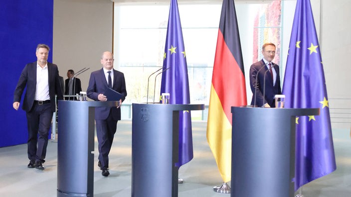 Bundeskanzler Olaf Scholz, Bundeswirtschaftsminister Robert Habeck, Bundesfinanzminister Christian Lindner gehen zu den Rednerpults. Im Hintergrund sieht man die Flaggen der Bundesrepublik und der EU.