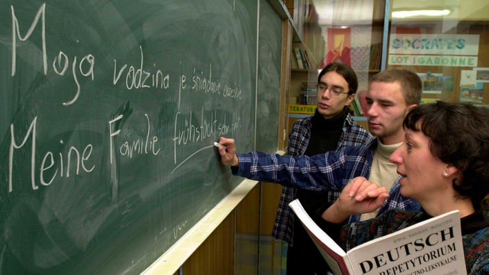 Symbolbild: Eine Lehrerin mit zwei älteren Schülern an der Tafel, an dem ein Satz auf polnisch und dessen deutsche Übersetzung steht.