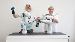 Frank H. P. Fitzek von der Technischen Universität Dresden, steht mit Datenhandschuhen neben Markus Rindt, Intendant der Dresdner Sinfoniker, an zwei Robotern, die ein Orchester dirigieren sollen.