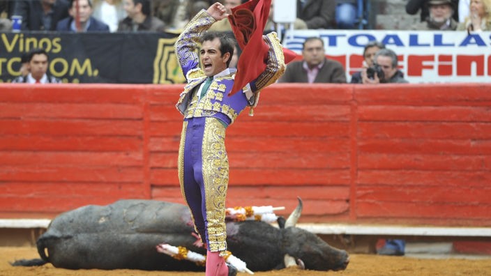 Ein Stierkämpfer feiert in der Arena seinen Sieg, indem er sein rotes Tuch in die Höhe hält. Am Boden liegt ein regungsloser Stier.