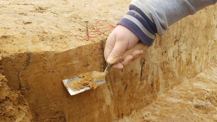 Zu sehen ist die Hand eines Mannes, welche eine Archäologenkelle bei einer Ausgrabung hält.