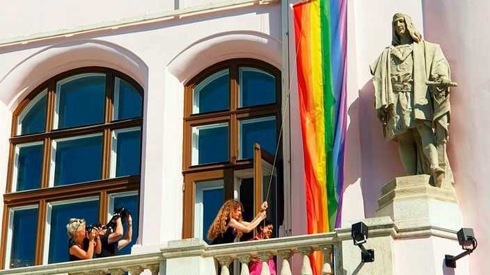 Direktorin Lotte de Beer hisst eine Regenbogen Flagen an der Front der Volksoper neben einer Statue.