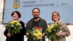 Drei Preisträger stehen mit Blumensträußen in der Hand auf der Bühne.