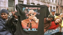 Fans entlang einer Absperrung; Ein Mann hält ein T-Shirt mit der Aufschrift "In memory of Notorius B.I.G." und dessen Gesischt hoch